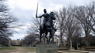 A statue of a woman wearing armor astride a horse.  She holds a sword in the air. 