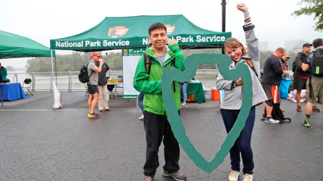 Two people hold a wooden arrowhead cut out in front of a large green pop up tent during an event