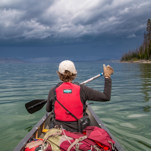 a boy in a red lifejacket paddles a canoe on a lake