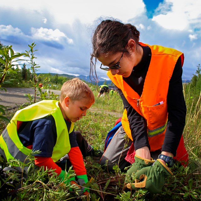 a young boy and girl in safety vests pull weeds