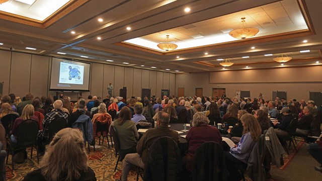People are sitting at round tables in a conference room, listening to a presenter