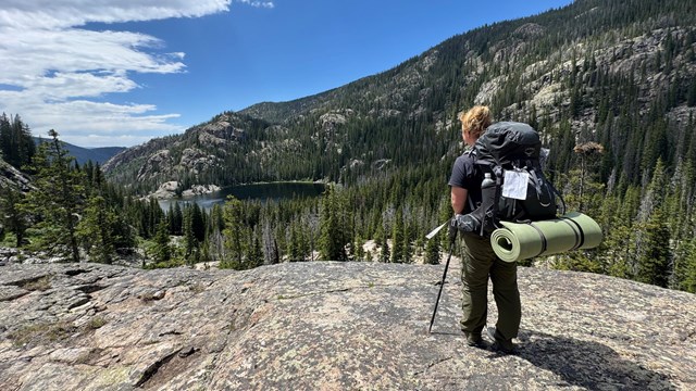 A person carrying an overnight backpack and permit is gazing out over a lake while on a trail