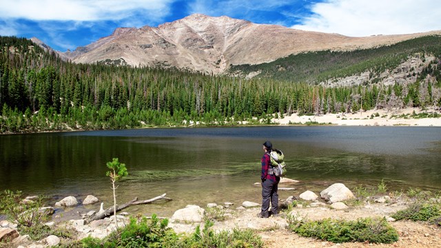 A person is standing at the edge of an alpine lake, carrying a backpack, on sunny day.