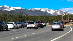 Cars waiting in line on roadway with snowy mountains in the distance.