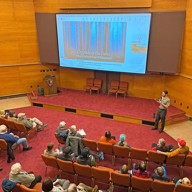 A park ranger is leading a talk in an auditorium with a slide presentation.