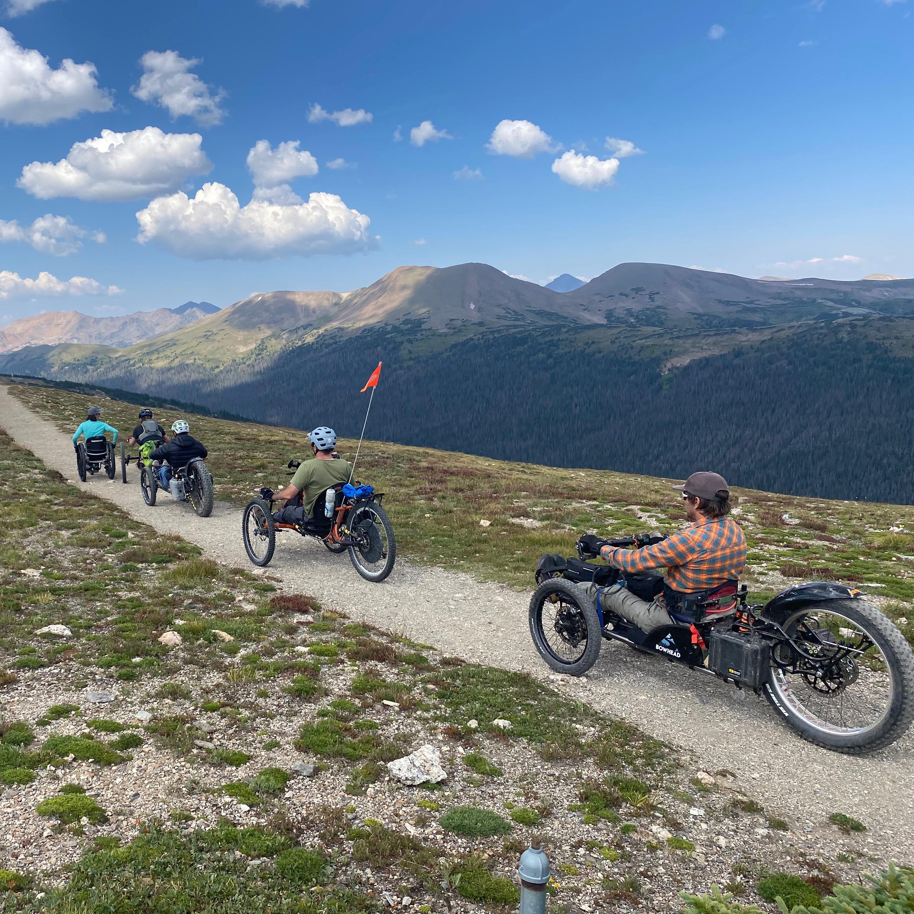 Five people in adaptive wheelchairs on a path through the tundra with large mountains behind them.