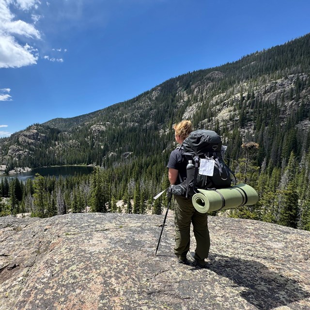 A person carrying an overnight backpack and permit is gazing out over a lake while on a trail