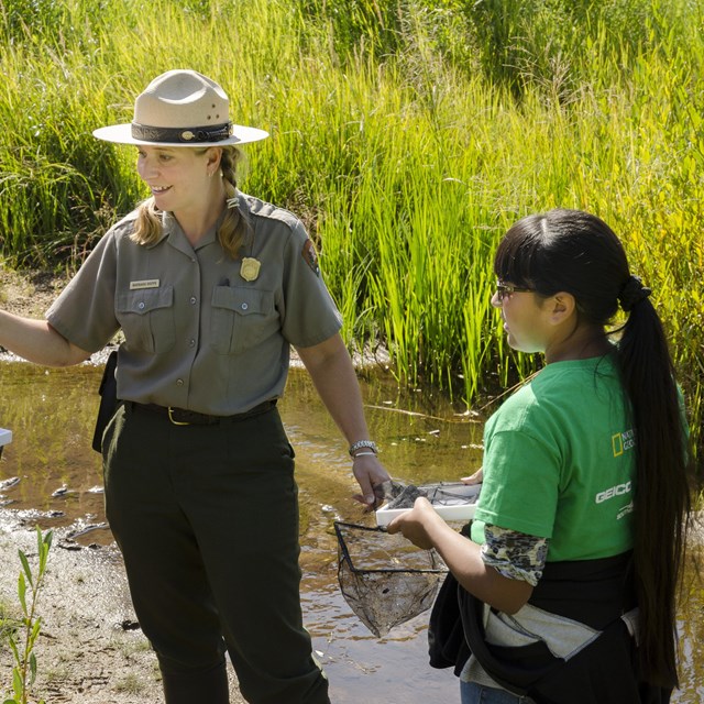 A park ranger is in a meadow leading a program with two youth