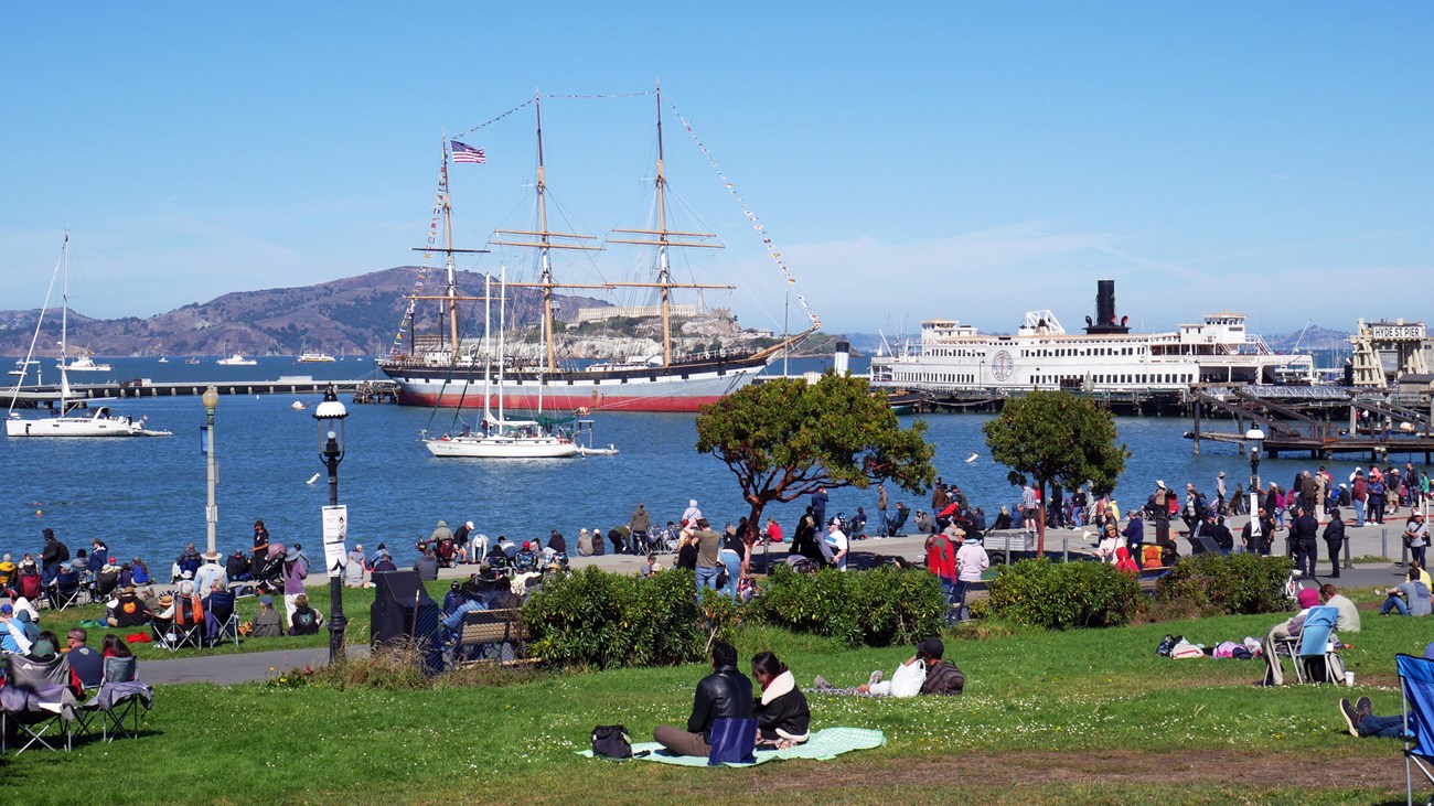 People sit on a grassy lawn with Aquatic Park Cove and Balclutha in the background