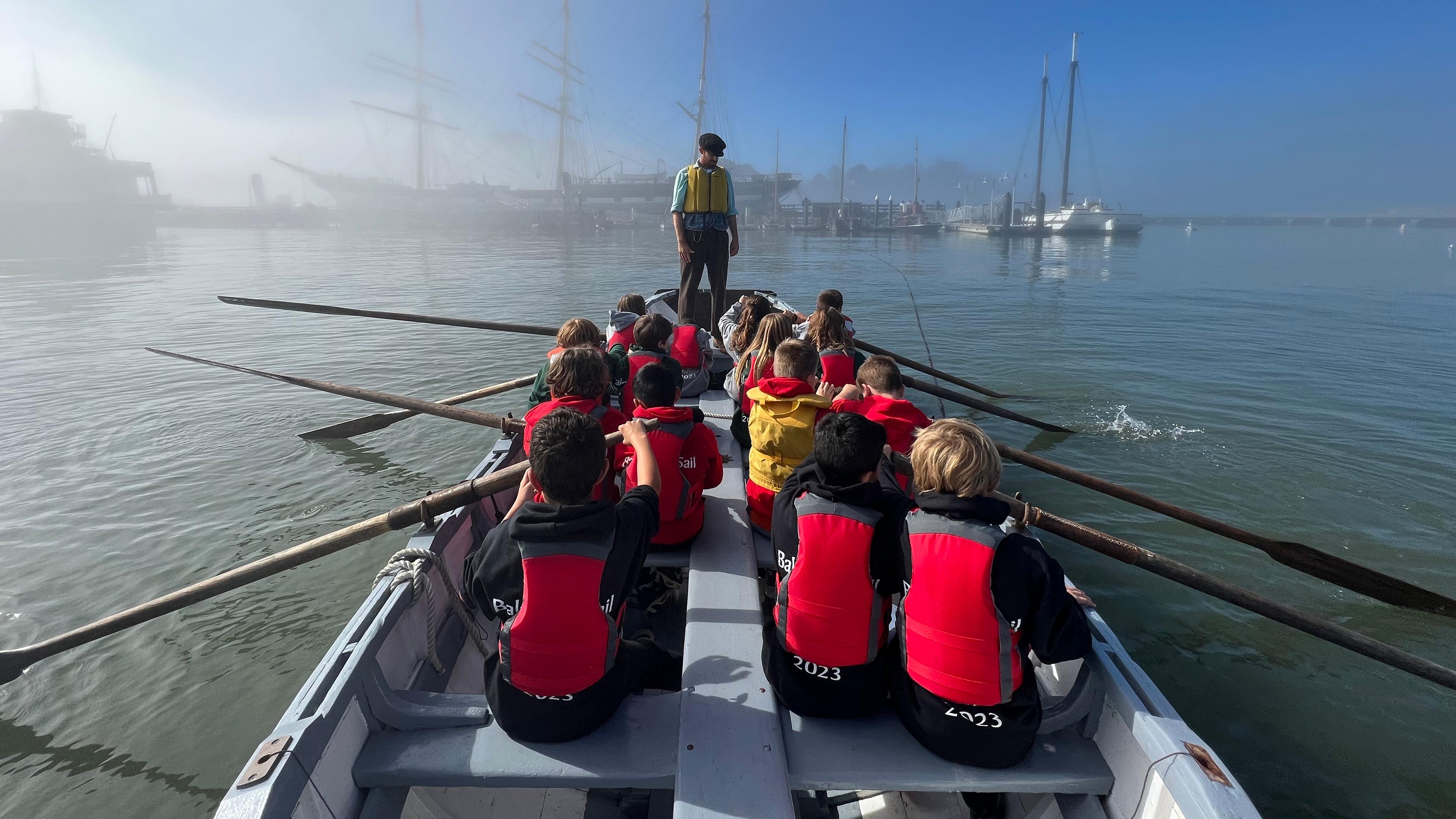 Children wearing life jackets sitting in a large row boat while an adult stands at the bow