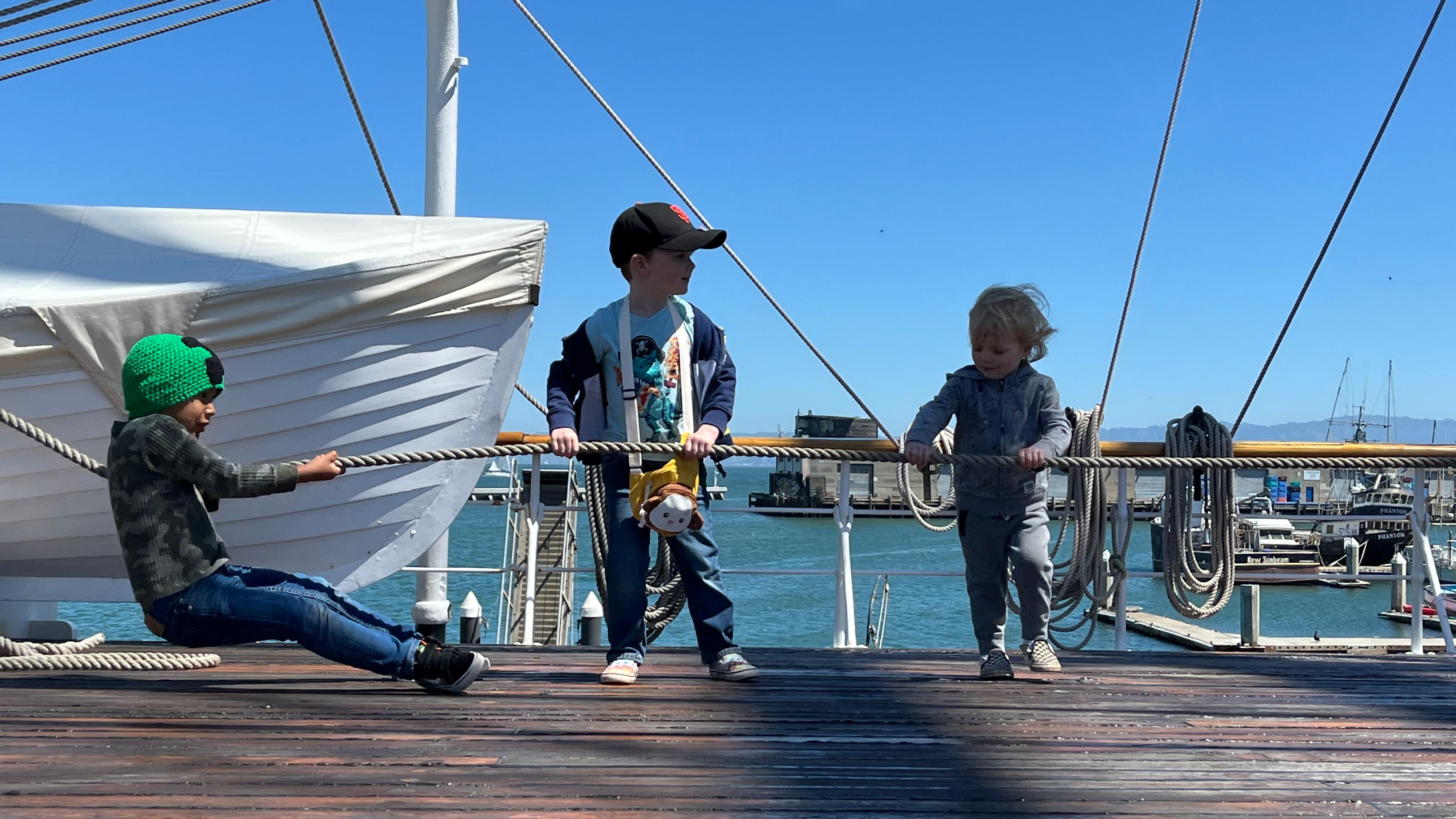 Three young children pull on a rope line on a ship deck.