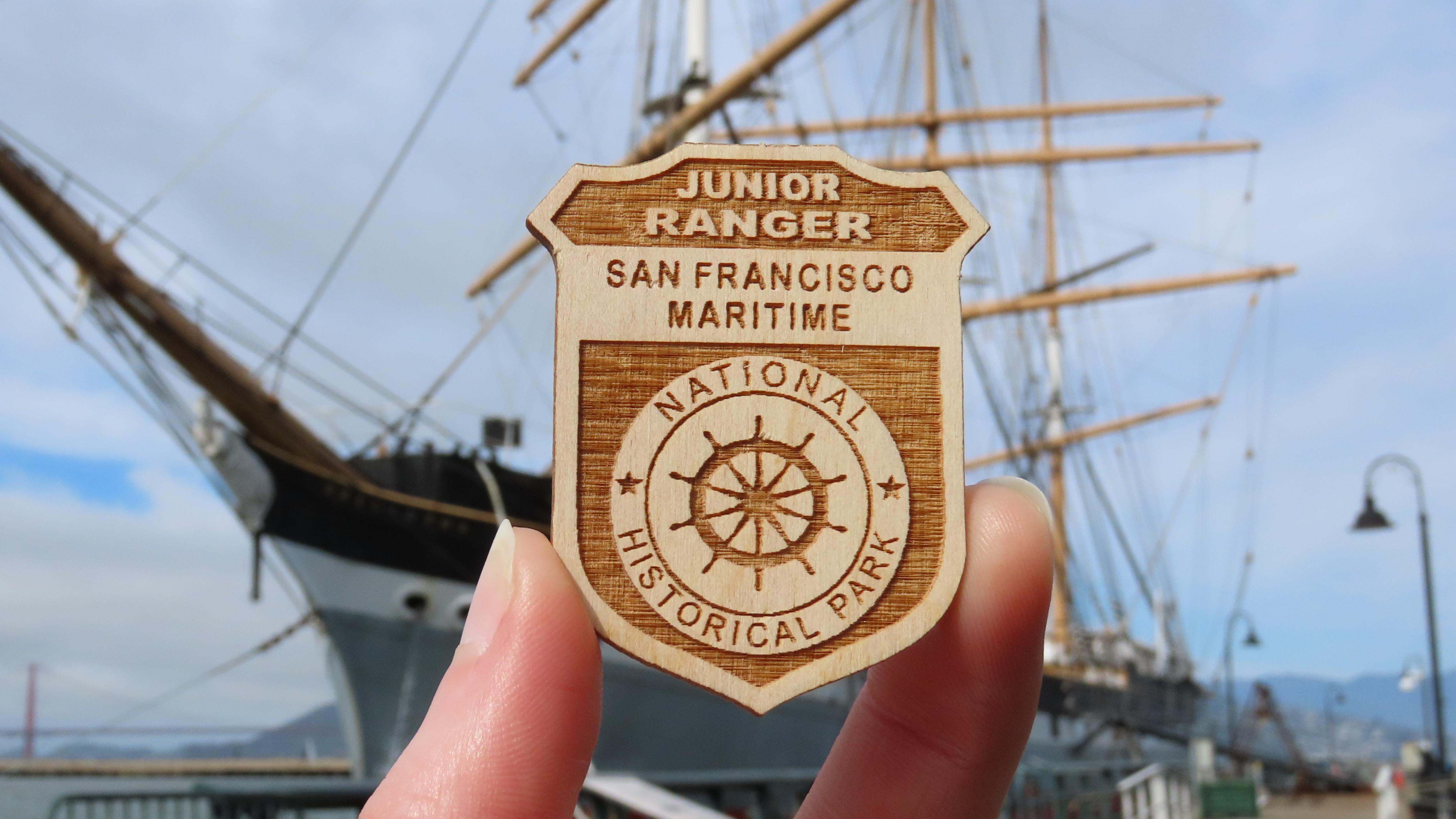 a San Francisco Maritime Junior Ranger Badge is held up, with a ship visible in the background.