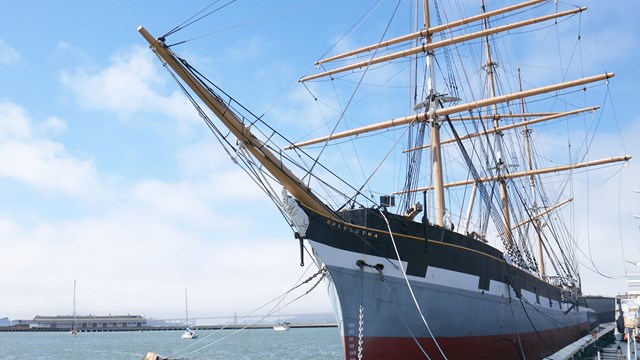 a three-masted sailing ship is tied to a pier, with the golden gate bridge in the distance