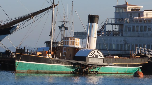 a medium-sized steamship with large side wheels and a green hull is docked to a pier