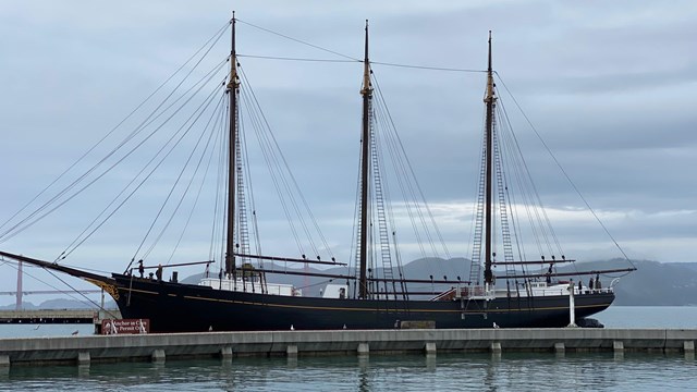 A large black wooden ship with three masts is docked to a pier