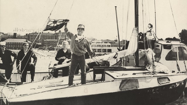 a young Japanese Man standing on the deck of a sailboat waiving a Japanese flag