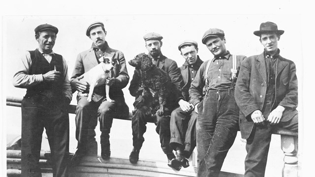 black and white photo from the early 20th century of men leaning against a railing, two holding dogs