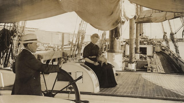 historic photo circa 1906 of a captain at the wheel of a ship and woman with a small dog on her lap