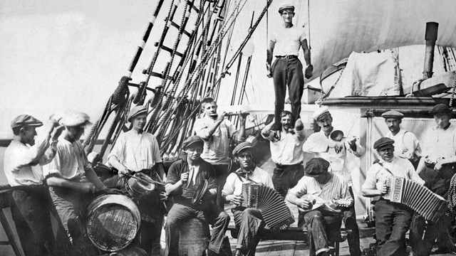 A group o sailors on the deck of sailing ship, seated and standing, holding musical instruments