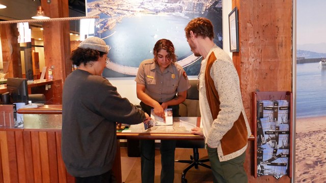 A park ranger at a visitor center desk points out directions on a map to two visitors