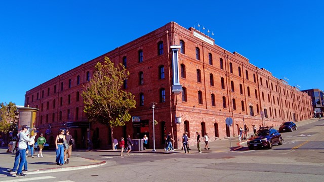 a historic brick warehouse stands on the corner of a busy street on a sunny day.