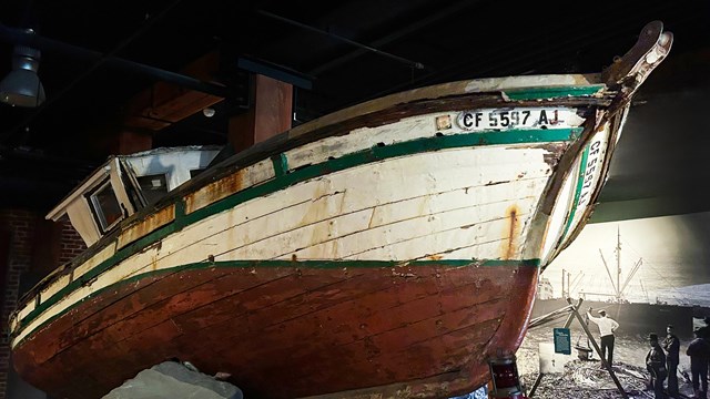 a wooden ship is displayed as a shipwreck exhibit in a dark hall with dramatic lighting. 