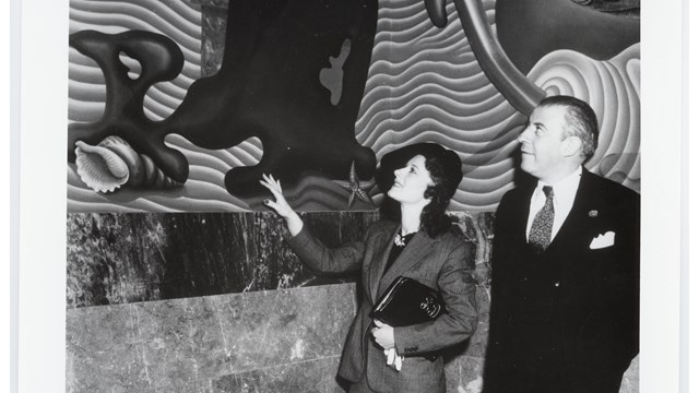 black and white image of Hilaire Hiler and a woman standing next to and looking up at a large mural