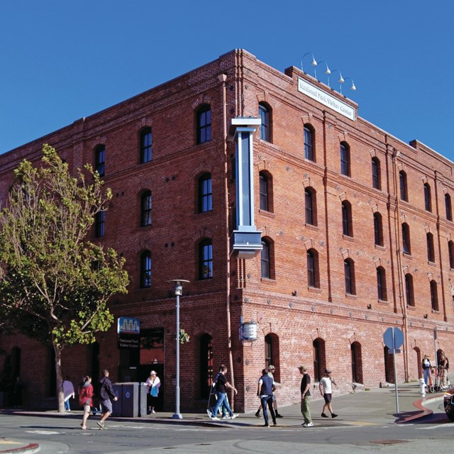 A historical brick warehouse stands on the corner of a busy street on a sunny day.