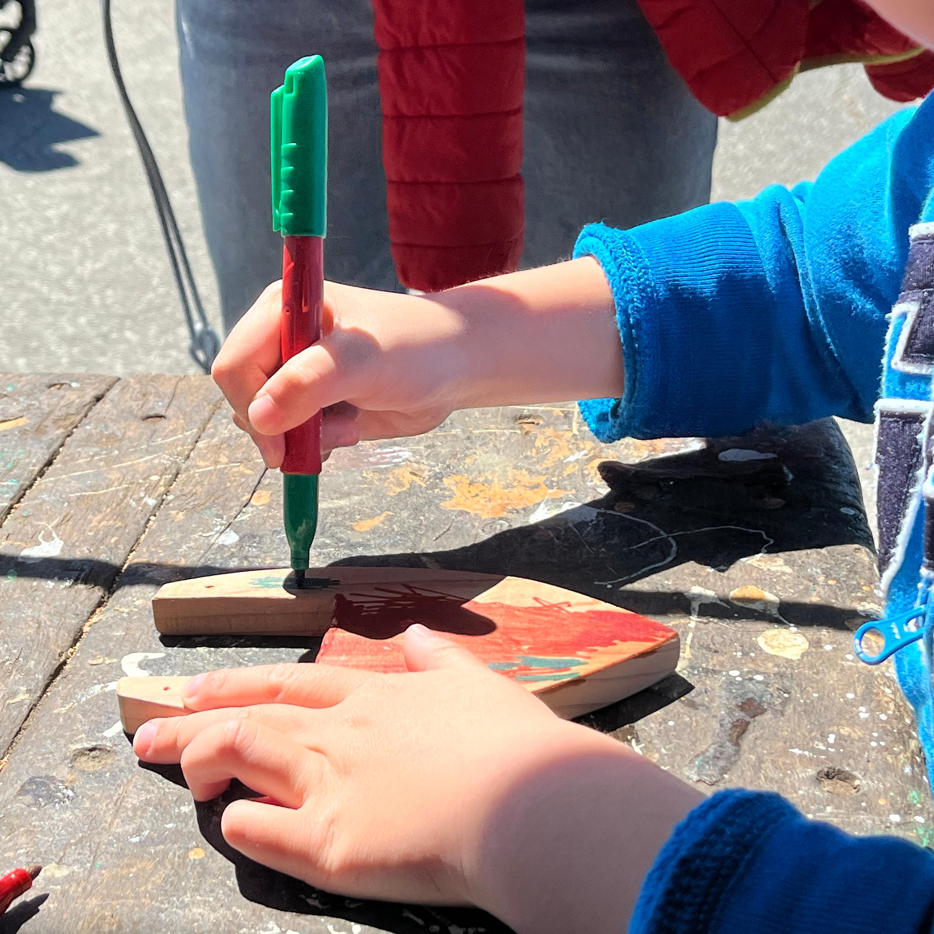 close-up of a child's hands drawing with a marker on a piece of shaped wood