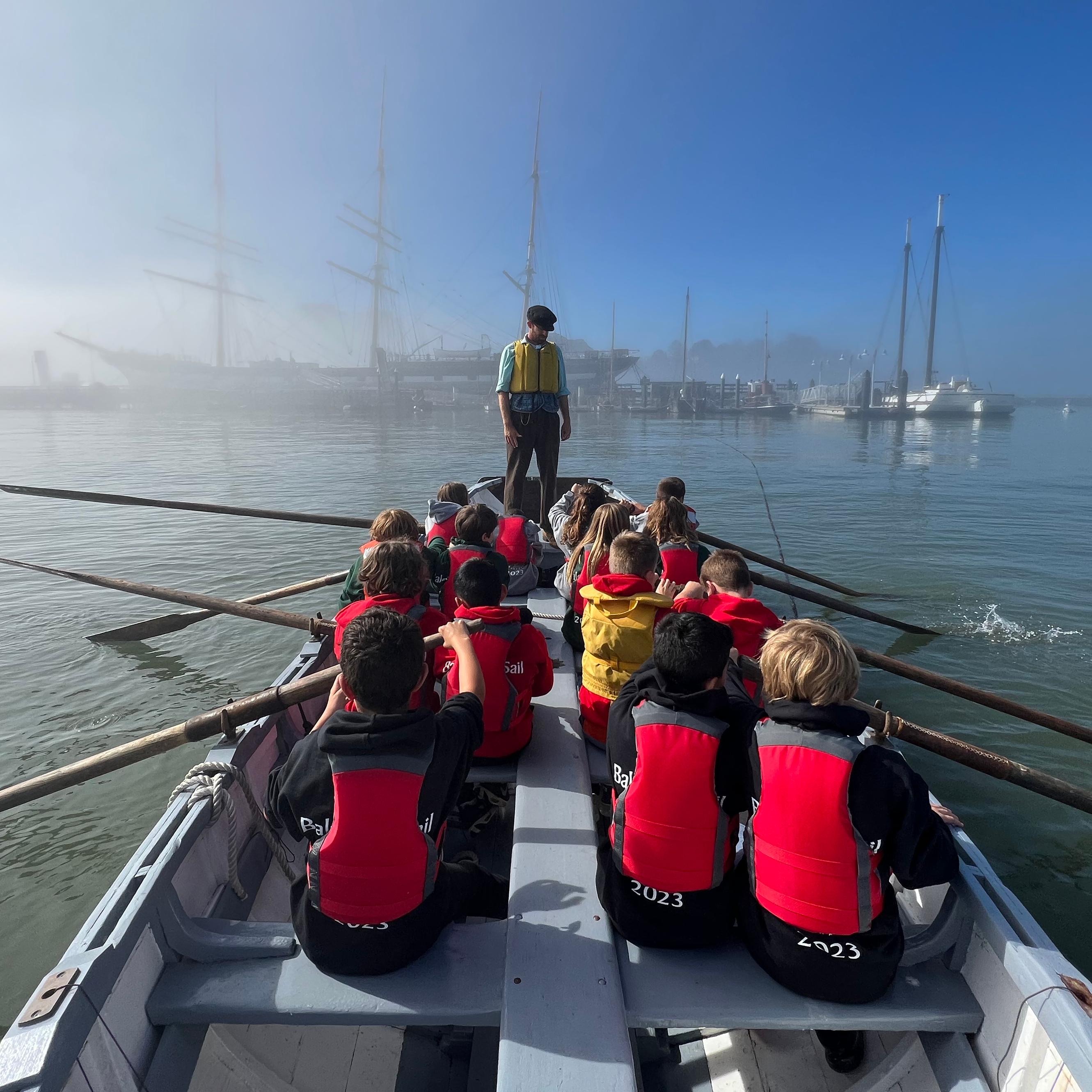Children wearing life jackets sitting in a large row boat while an adult stands at the bow