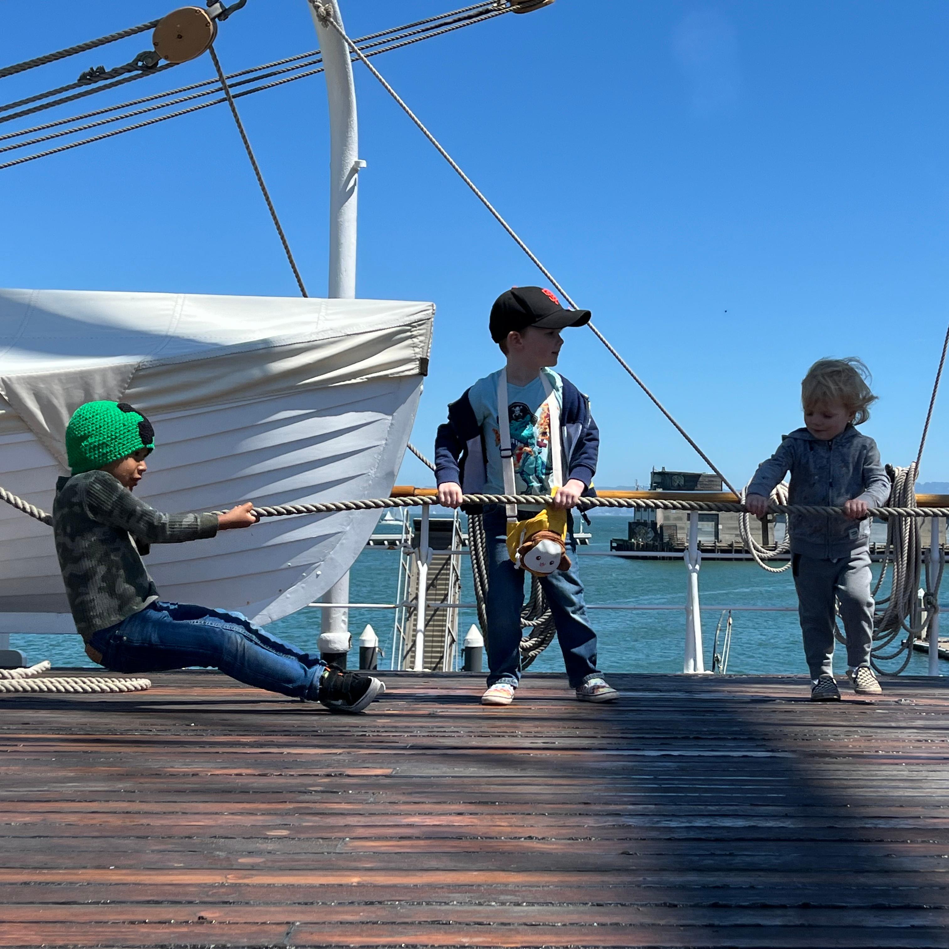 Three young children pull on a rope line on a ship deck.