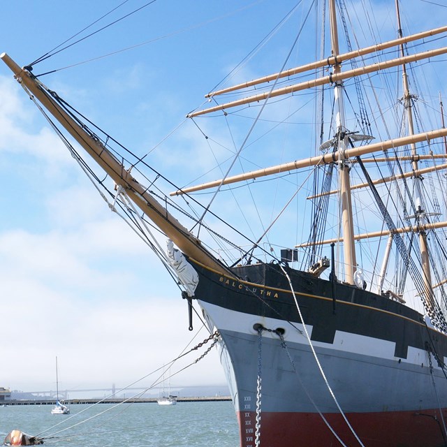 a three-masted sailing ship is tied to a pier, with the golden gate bridge in the distance