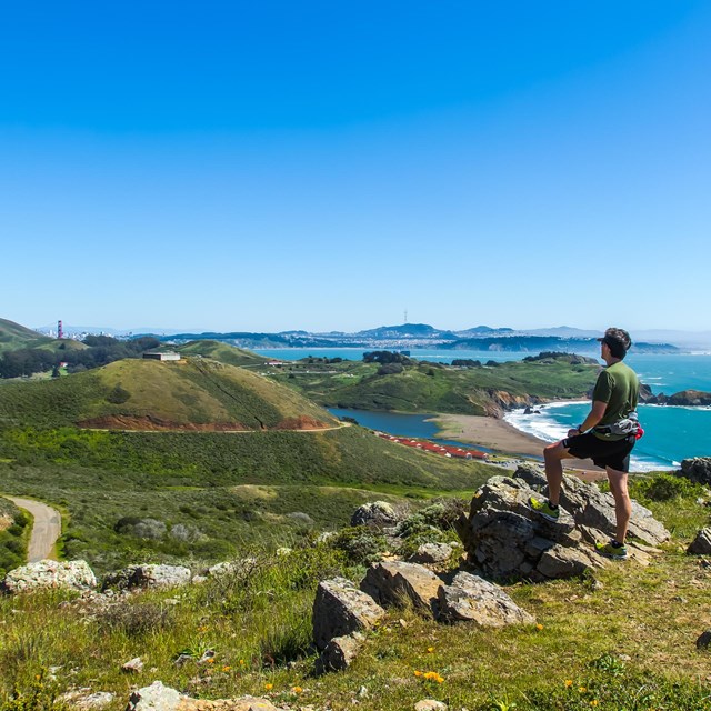 a hiker stands on a rocky hill overlooking the golden gate bridge in the distance