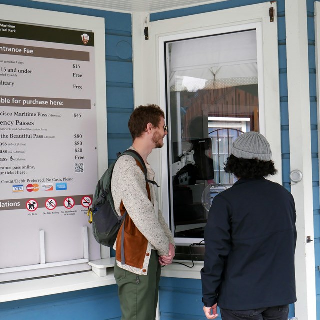 Two visitors stand at a fee booth window next to a sign with fee and pass information