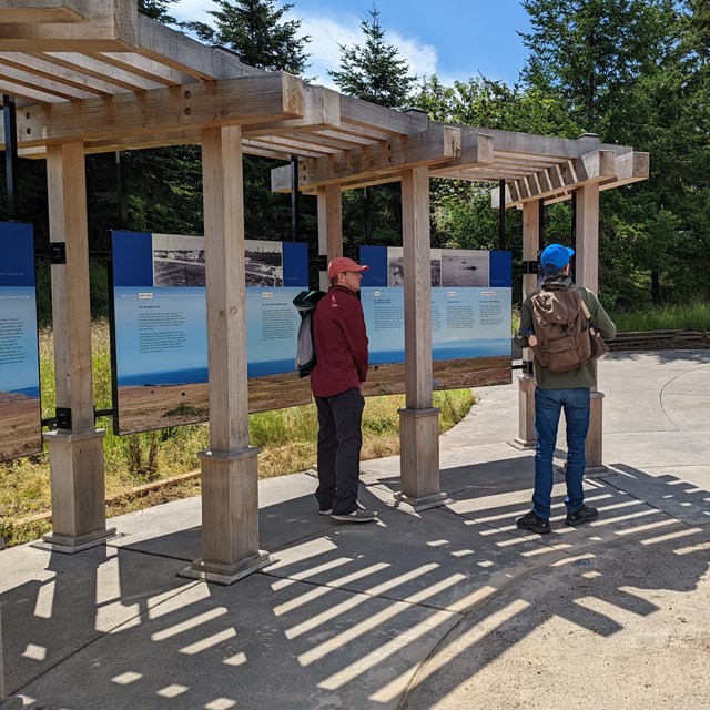 Three visitors stand outside of the new visitors center. Educational signs in the background