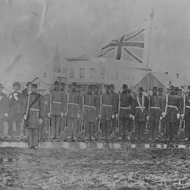 Black and white photograph of men in british uniform drilling in a field with a uk flag behind them