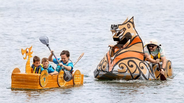 Two groups in decorated cardboard boats race in a harbor