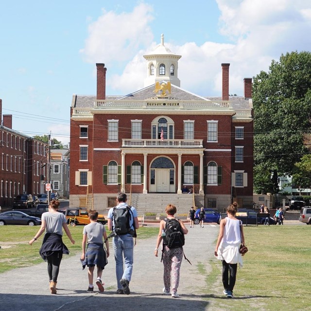 A group of five people walk toward a brick building.