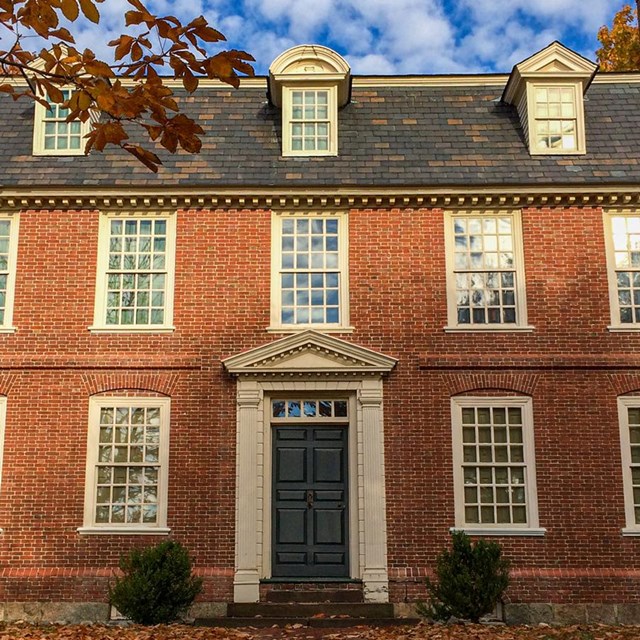 Two story brick house with many windows and leaves on the ground indicate it is fall