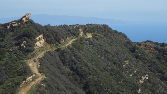 Hikers in the distance hiking along a mountain trail with the pacific ocean in the background.