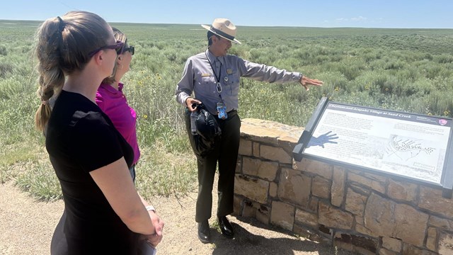 A park ranger gestures at an exhibit panel in a prairie landscape. 
