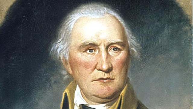 An older man in an 18th century uniform with white shirt and blue jacket with gold details.