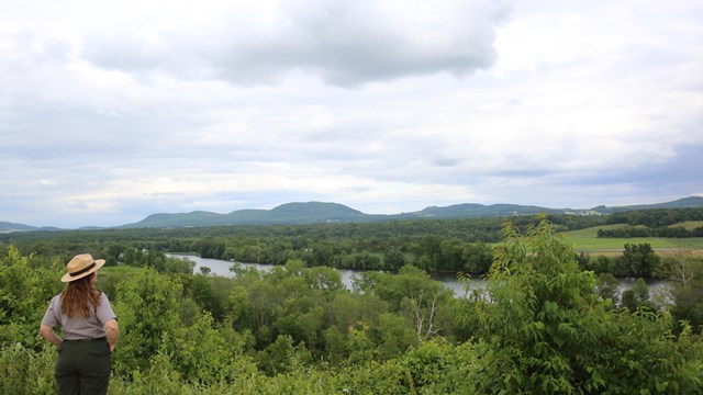 A woman in a ranger uniform stands near an overlook of the Hudson River with green vegetation.