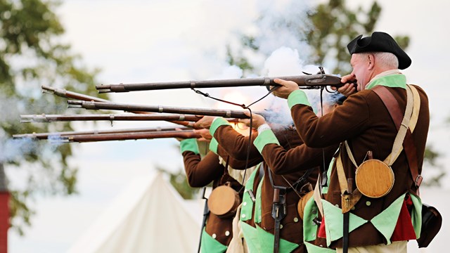 18th Century Soldiers in brown and green uniforms fire muskets.