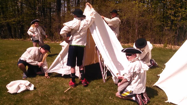 Students in white 18th century style shirts work together to pitch a canvas tent. 