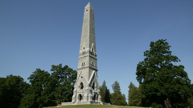 A clear blue sky over a tall, gray obelisk monument on a small hill. 