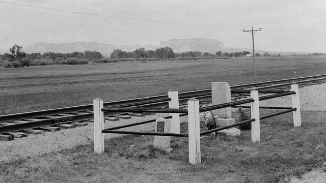 A black and white photo of a headstone, surrounded by a pipe fence, located next to railroad tracks.