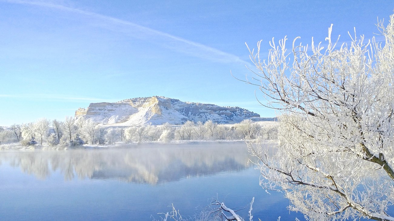 A snow-covered bluff is reflected in the water of a pond. 