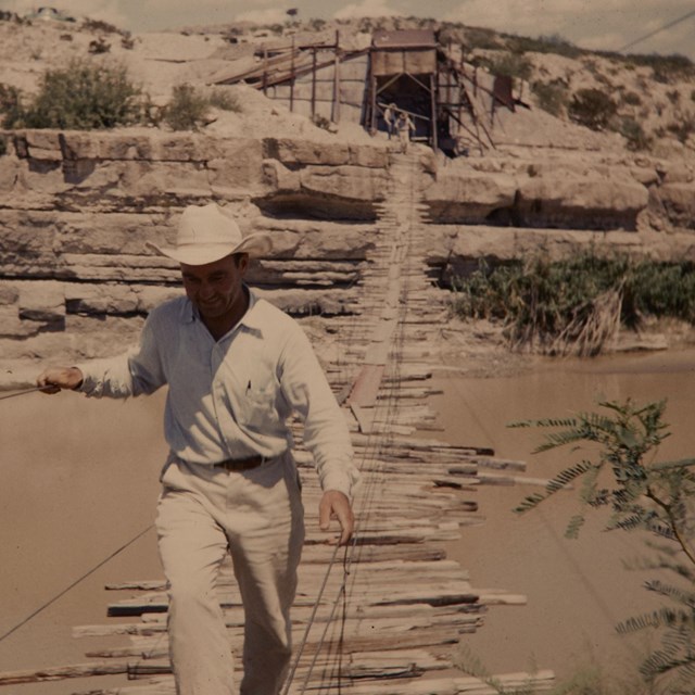 Man walking across wooden plank bridge over Rio Grande River with two buildings behind it.