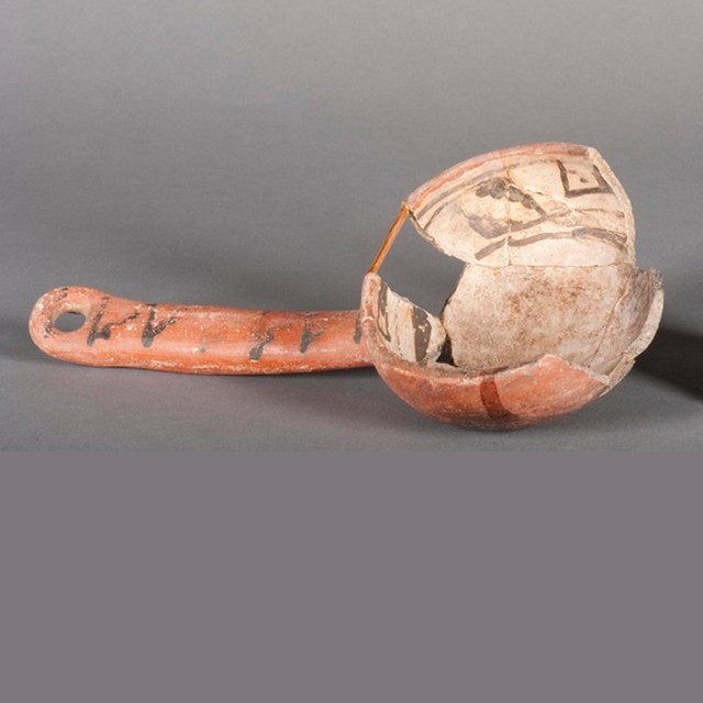 A pottery ladle, orangey in color, with designs on the inside; pieces missing from the bowl.